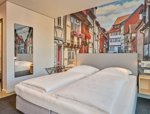 Modernes Doppelzimmer mit großem Bett, hellem Design und Fotowand historischer Fachwerkhäuser.