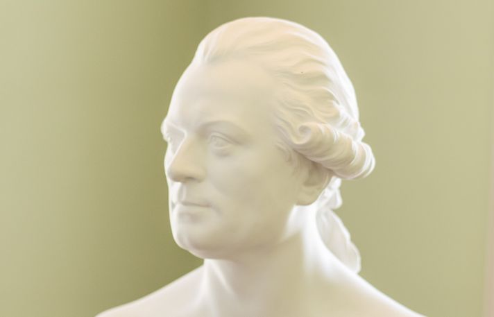Weiße Marmorbüste eines Mannes mit seitlich gewelltem Haar vor olivgrünem Hintergrund