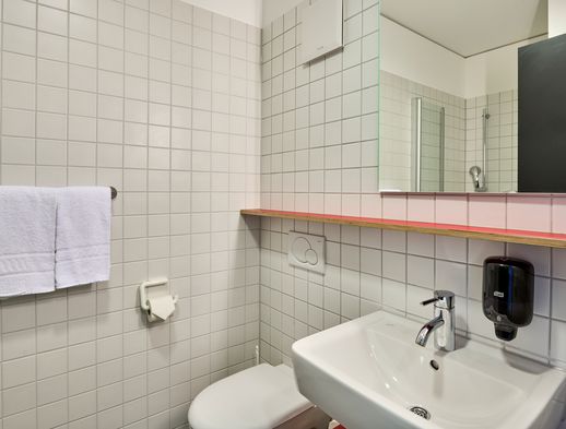 Helles Badezimmer mit weiß gefliesten Wänden, Waschbecken, Spiegel, Handtuch und Seifenspender.