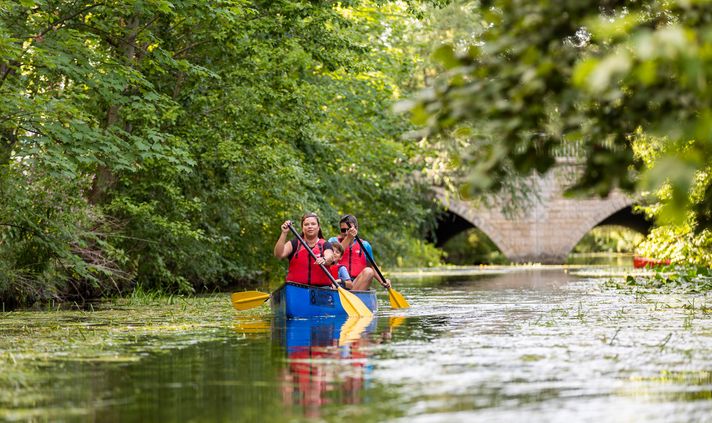 Zwei Erwachsene und ein Kind paddeln im Kanu auf einem grünen Fluss unter einer Steinbrücke.